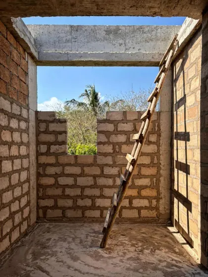 A simple wooden ladder leans against a wall in a room under construction at Kizikula. The image captures the raw, handmade quality of the building process, with sunlight streaming through the open roof.