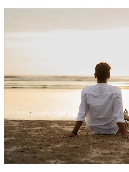 A contemplative pre-wedding portrait of the groom-to-be sitting on the beach, looking out at the ocean at sunset.
