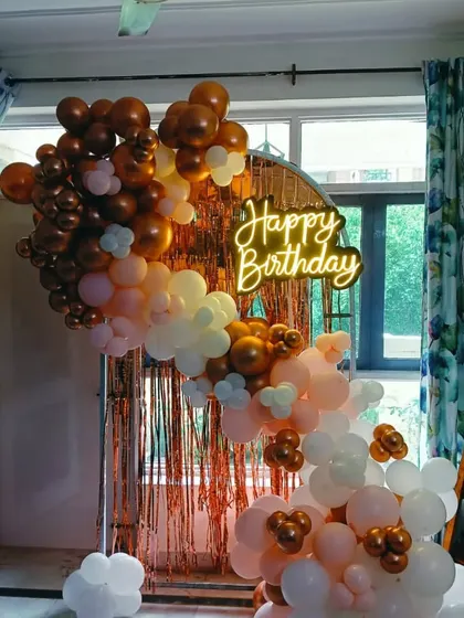 A birthday decoration with a rose gold fringe backdrop. The setup features a balloon arch in bronze, peach, and white, and a "Happy Birthday" neon sign.