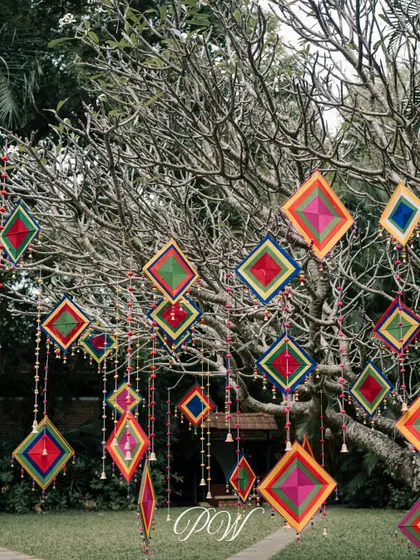 Colorful kite-like hangings and bells adorn a tree for a Mehendi ceremony, adding a playful and festive touch to the outdoor space.