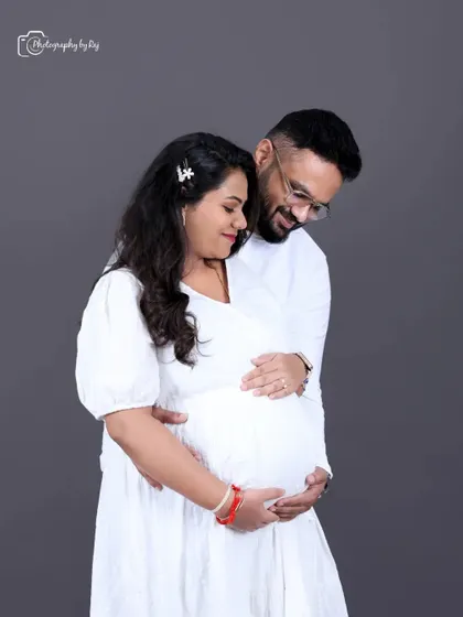 A simple and sweet couple's portrait in matching white outfits against a solid grey backdrop. The focus is purely on their connection and anticipation.