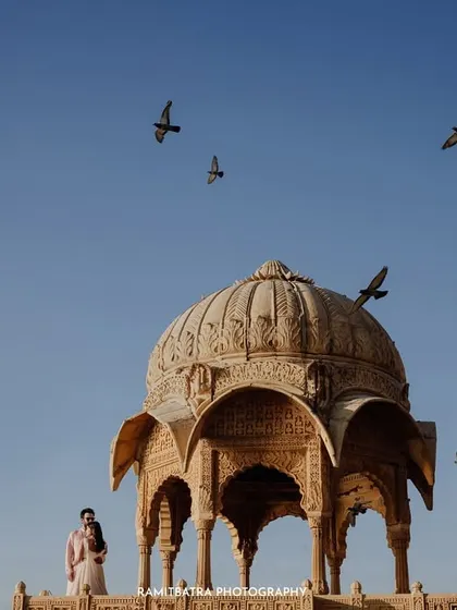 The couple stands before a historic cenotaph in Jaisalmer as pigeons take flight, creating a dynamic and poetic frame.