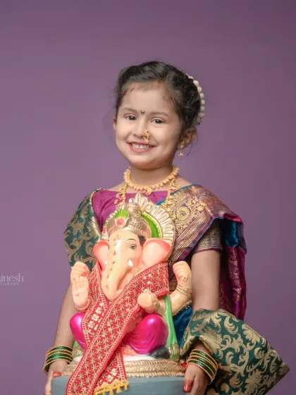A close-up portrait of a little girl in a traditional saree, holding the Ganpati idol. Her sweet smile and authentic attire make this a heartwarming festival photograph.