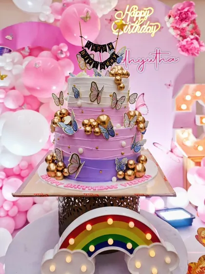 A close-up of a stunning three-tiered birthday cake adorned with butterflies, set against a pink backdrop with a 'Happy Birthday' neon sign and marquee number.