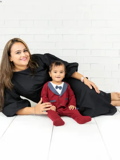 A mother lies comfortably on the floor next to her baby boy, who is dressed smartly in a little tuxedo-style onesie. A sweet and stylish mother-son portrait.