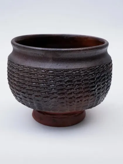 A dark, textured bowl with a woven pattern carved into the clay. The deep brown glaze breaks over the texture, highlighting the details and giving it a rustic, tactile quality.