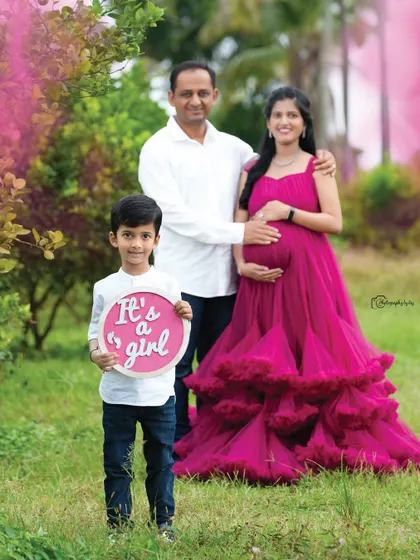 A joyous gender reveal moment for a growing family. The older brother proudly holds an "It's a girl" sign, while his parents stand behind him, sharing a happy embrace in our beautiful outdoor location. We provide the props to make your announcement special.