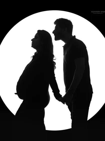 A classic couple's silhouette against a circular light source. This timeless black and white shot emphasizes their bond and the shape of the baby bump.