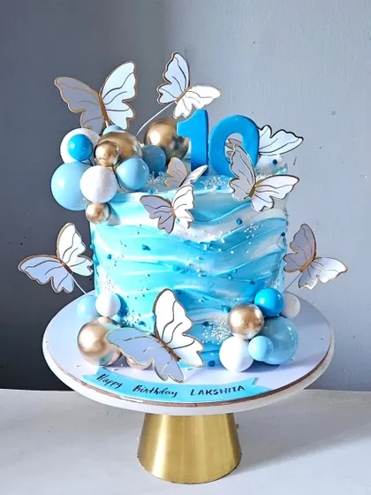 A beautiful butterfly-themed cake in shades of blue. This design features a watercolor effect on the cream, adorned with delicate paper butterflies and decorative spheres for a light and airy feel.