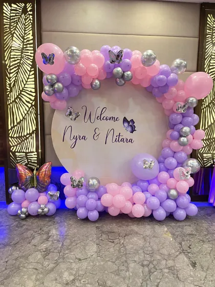 A welcome decoration with a butterfly theme. This setup features a circular backdrop with custom names and a balloon arch in purple and pink.