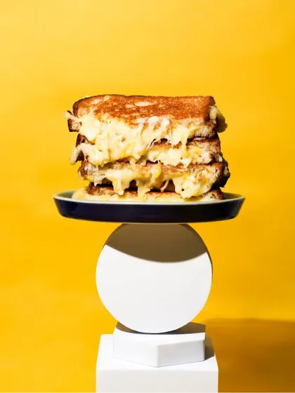 A towering stack of gooey grilled cheese sandwiches, balanced on geometric blocks against a bold yellow background. This quirky and minimalist shot is designed to be eye-catching and memorable.