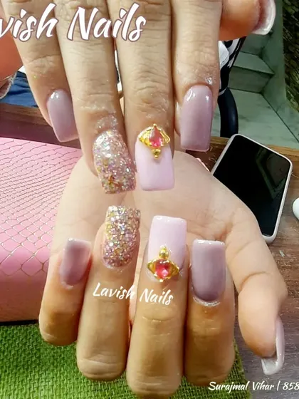 A unique design featuring a large, gold-encased pink gemstone as a centerpiece on a light pink nail, paired with glitter and solid-colored nails.