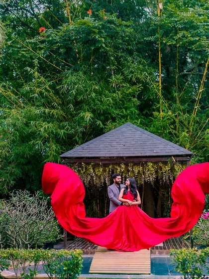 A wide shot showing the couple embraced at the gazebo. The flowing red gown creates a perfect semi-circle, beautifully framing them against the green background.