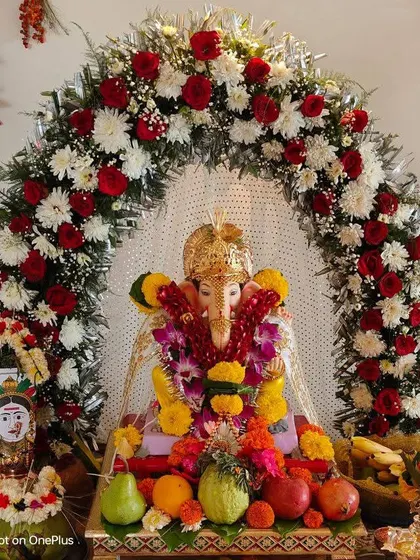 A floral arch made of red roses, white chrysanthemums, and greenery placed around a Ganesha idol for a puja. The base is decorated with fresh fruits and offerings.