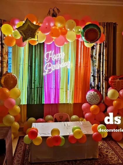 A vibrant and unique fruit-themed birthday party. This colorful setup uses a rainbow crepe paper backdrop, fruit-shaped foil balloons, and a central neon sign.
