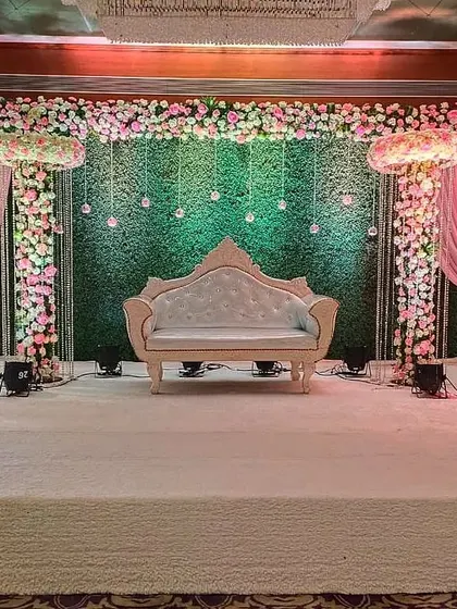 An extravagant reception stage with floral pillars, a lush green backdrop, and hanging light bulbs. The pink draping adds a soft, romantic touch to the overall grand design.