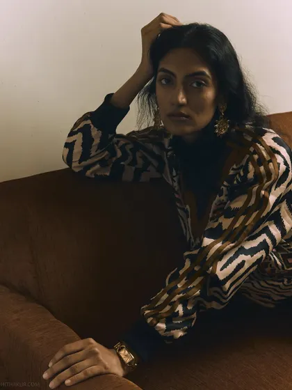 An editorial fashion shoot with a moody, intimate feel. The warm lighting and relaxed pose on the sofa create a comfortable yet stylish atmosphere.