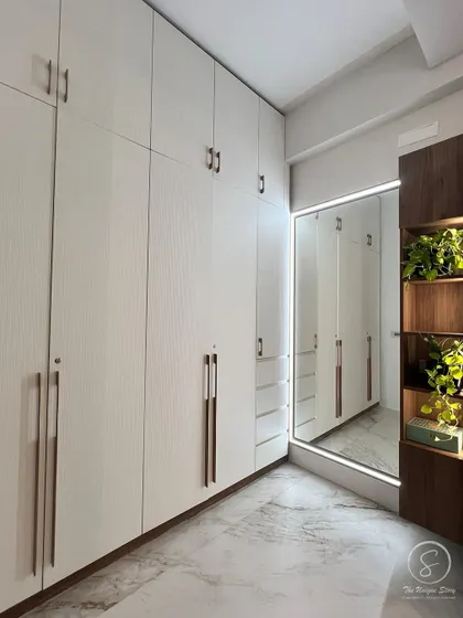 A walk-in closet with clean, white wardrobes and minimalist handles. An open wooden shelving unit with integrated lighting provides a warm contrast and a perfect spot for displaying plants and personal items.