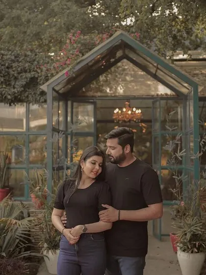 A romantic pre-wedding portrait of a couple in a lush garden, standing before a beautiful greenhouse, capturing a moment of quiet connection.