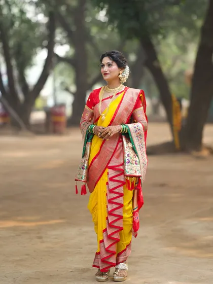 A stunning full-length shot of a bride in a traditional yellow and red Nauvari saree. The look is completed with an elegant updo, gajra, and classic gold jewelry, perfect for a rustic-themed wedding shoot.