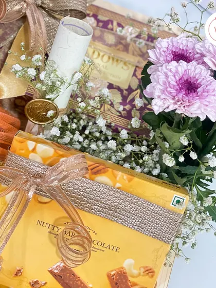 This close-up shows the details within the wooden tray hamper. You can see the hand-rolled invitation scroll, sealed with a custom wax seal, nestled beside nutty chocolate bars and fresh flowers.