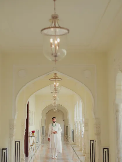 The groom walks down a grand, sunlit corridor of a palace, the repeating arches creating a sense of scale and majesty.