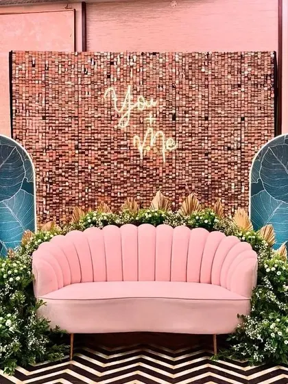 A chic and modern photo booth for a party. It features a rose gold sequin wall, a "You & Me" neon sign, and tropical leaf panels, all centered around a stylish pink sofa.