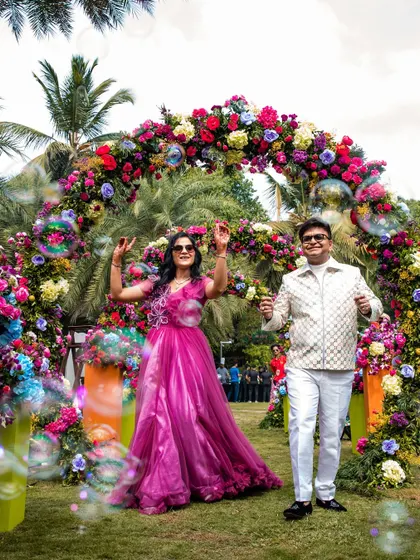 A couple making a grand entrance through a vibrant floral arch with bubbles, celebrating their 25th anniversary in style at W Goa.