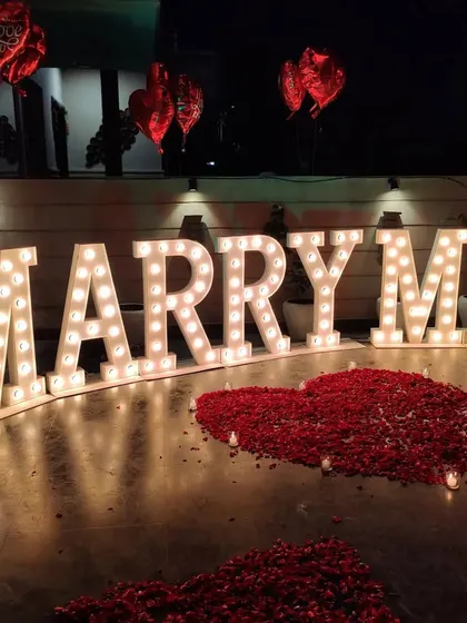 Another angle of our terrace proposal setup, showing the glowing marquee letters and heart-shaped arrangements of red rose petals. Red foil heart balloons add a final romantic touch to the scene.