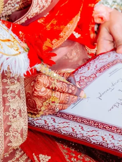 A close-up shot of the bride's henna-adorned hand as she signs the Nikah nama. The decorated pen and the intricate border of the certificate add to the beauty of the moment.