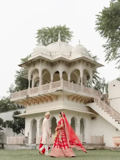 The couple takes a walk in the palace gardens, the bride's red lehenga flowing beautifully. This wide shot captures the romance of the moment and the grandeur of the location.