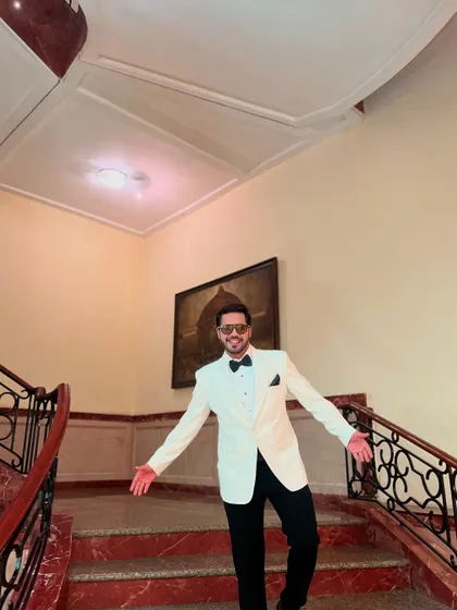 A welcoming, open-armed pose on a grand staircase, dressed in a sharp white tuxedo.