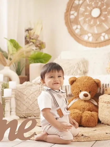 A charming first birthday portrait of a baby boy in a boho-chic setup, featuring a large wooden 'One' sign and a friendly teddy bear.