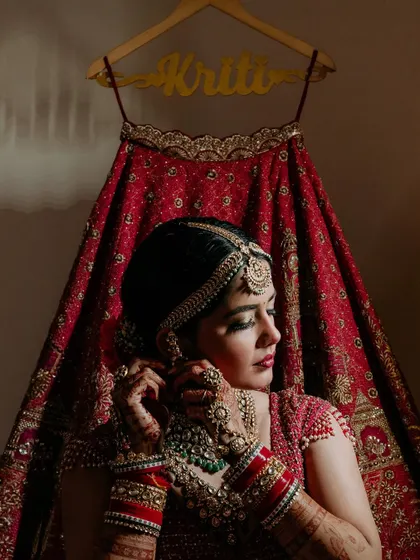 The final touches. A beautiful shot of the bride adjusting her earring, with her personalized wedding lehenga hanging in the background, telling a complete story in one frame.