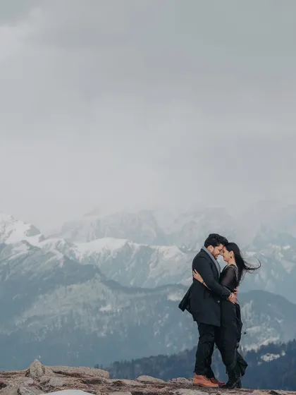 An epic wide shot of a couple embracing on a mountain peak. The wind catching her hair and the vast, moody sky create a dramatic and cinematic feel.