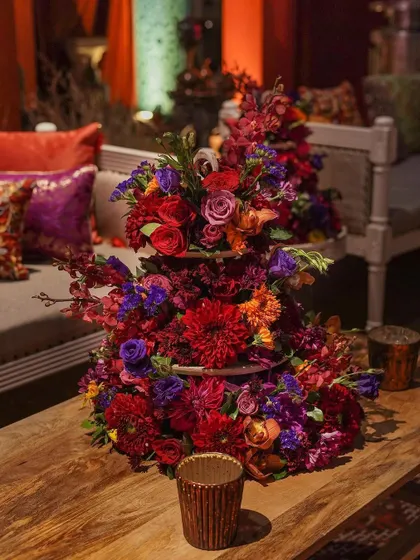 A tiered floral arrangement in deep reds and purples sits on a wooden table, echoing the rich color palette of the Moroccan theme. This centerpiece adds a touch of floral artistry to the rustic setting.