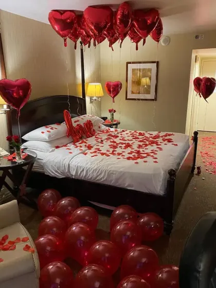 The ultimate romantic gesture with a classic red theme. This hotel room is adorned with red heart-shaped foil balloons, a "Love" balloon on the bed, and a floor scattered with rose petals.