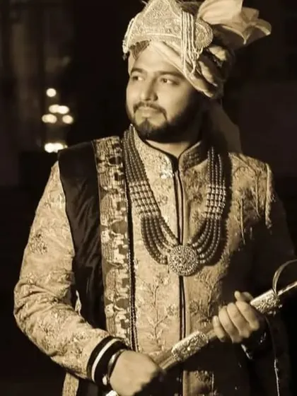 A classic and regal portrait of the groom in his wedding sherwani, holding a traditional sword. The sepia tone gives the image a vintage, timeless quality.