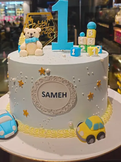 A charming cake for a baby boy's first birthday. This design is decorated with a fondant teddy bear, toy cars, and building blocks, celebrating one year of sweet moments.