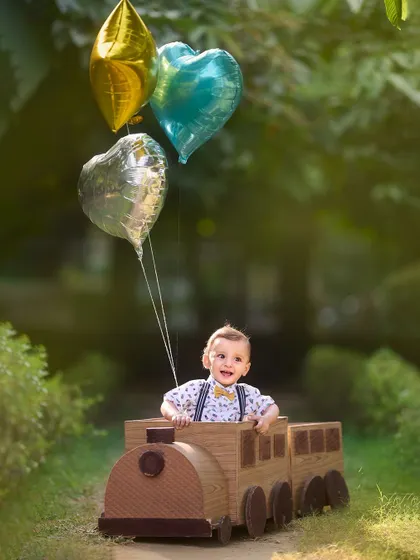 This little conductor is ready for an adventure. The toy train prop, complete with colorful balloons, adds a sense of fun and imagination to this outdoor baby photoshoot.