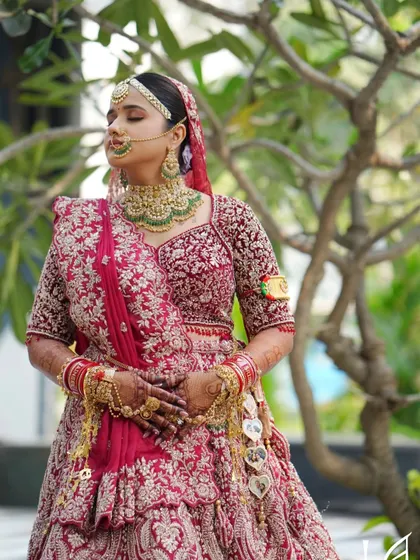 A serene outdoor portrait. The soft, natural light shows off the flawless skin work and the beautiful details of her red lehenga.