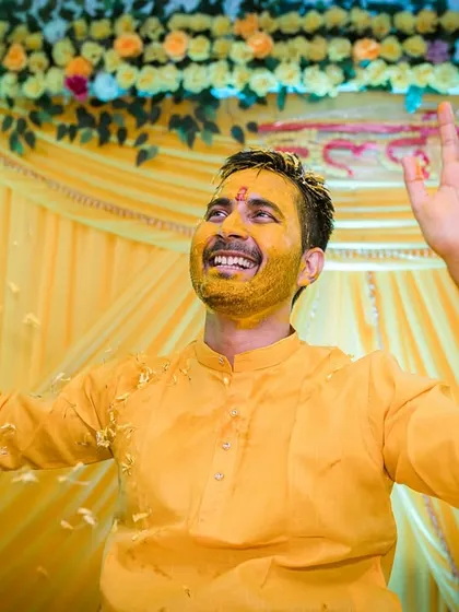 A moment of celebration. The groom, covered in turmeric and rice, raises his hands in the air, embracing the chaos and joy of the ritual.