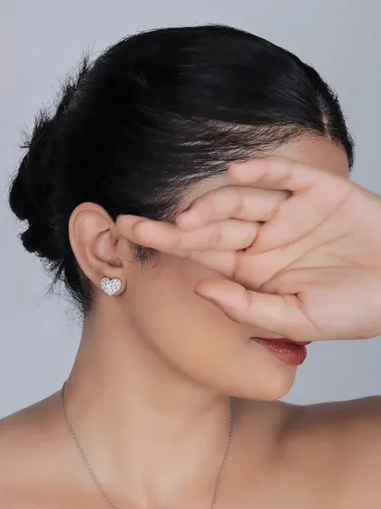 A minimalist lifestyle shot focusing on a single, elegant heart-shaped stud earring. The pose conceals the model's face, drawing all attention to the delicate piece of jewelry.