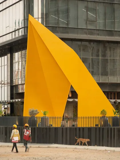 The "Yello" installation acts as a gateway and a landmark. Its bold color and folded geometric form create a striking contrast with the glass facade of the office building, giving the entrance a unique and memorable identity.