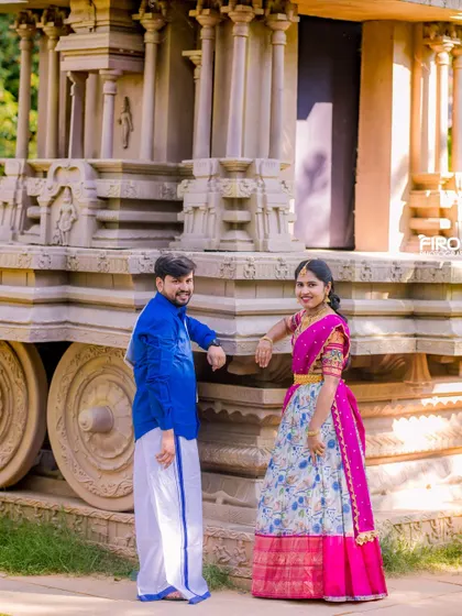 A stylish portrait of a couple in traditional South Indian attire by a stone chariot. The vibrant blue and pink outfits create a beautiful color combination.