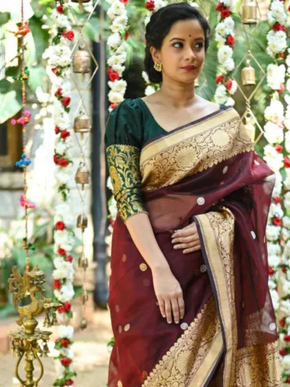 This rich maroon Kanjivaram silk saree is paired with a contrasting green brocade blouse with puff sleeves, creating a regal and traditional look.