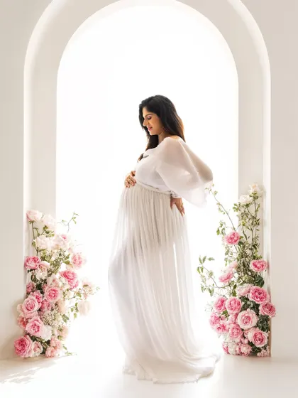 A side profile of the mom-to-be in the floral archway, cradling her bump. The combination of the white gown and pink flowers is simply perfect.