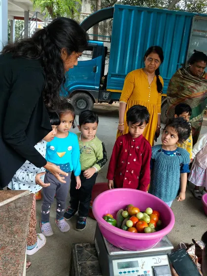 At the market, children observe a vendor weighing fresh tomatoes. This is a practical introduction to concepts of weight, measurement, and commerce in a real-world setting.