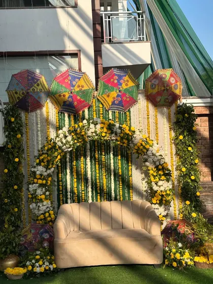 The full view of the sunny outdoor Haldi stage, featuring a comfortable sofa under a lush floral arch. The natural light brings out the vibrant colors of the decor.