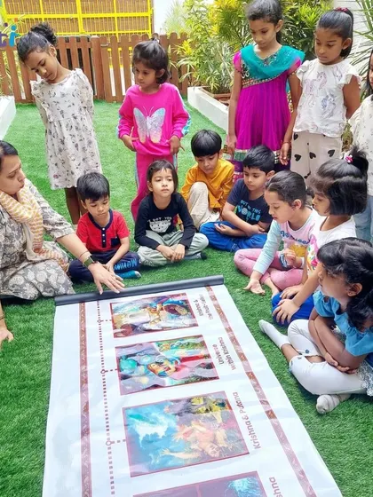 A teacher points to an illustration on a story scroll, explaining a scene from Krishna's life. Visual aids make the stories more engaging and memorable for young learners.